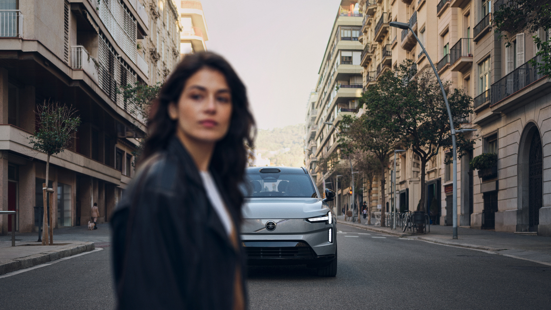 A young woman crosses a main road, positioned in the foreground out of focus. In the background is the new Volvo EX90, in focus, seemingly driving towards her.